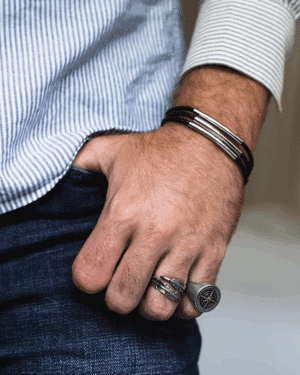 Muñeca masculina con pulsera de cuero y acero Valurys, anillos de acero quirúrgico con diseños decorativos, mano en bolsillo de pantalón vaquero.