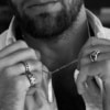 Hombre con camisa abierta sosteniendo colgante rectangular Valurys y luciendo anillos de acero en ambas manos.