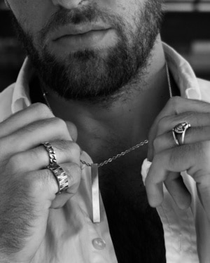 Hombre con camisa abierta sosteniendo colgante rectangular Valurys y luciendo anillos de acero en ambas manos.