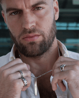 Hombre con barba sosteniendo el colgante Eclipse Valurys de acero quirúrgico, luciendo también anillos robustos y pulsera trenzada en la muñeca.