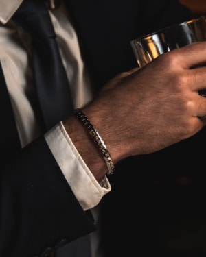 Muñeca masculina con pulsera de eslabones anchos de acero quirúrgico Valurys y camisa blanca, sosteniendo un vaso.